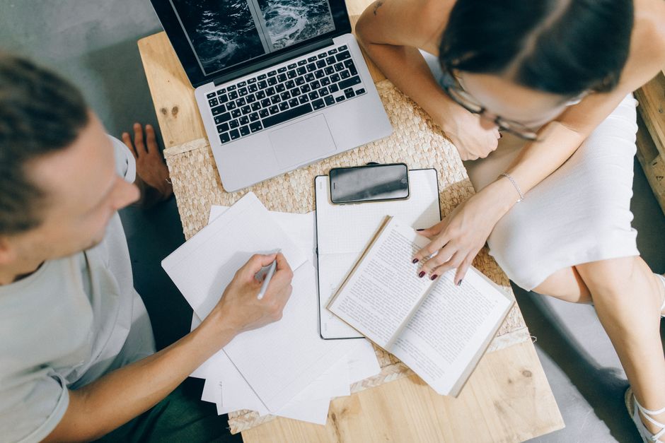 Overhead view of coworkers discussing ideas with laptop, papers, and books on a wooden desk.
